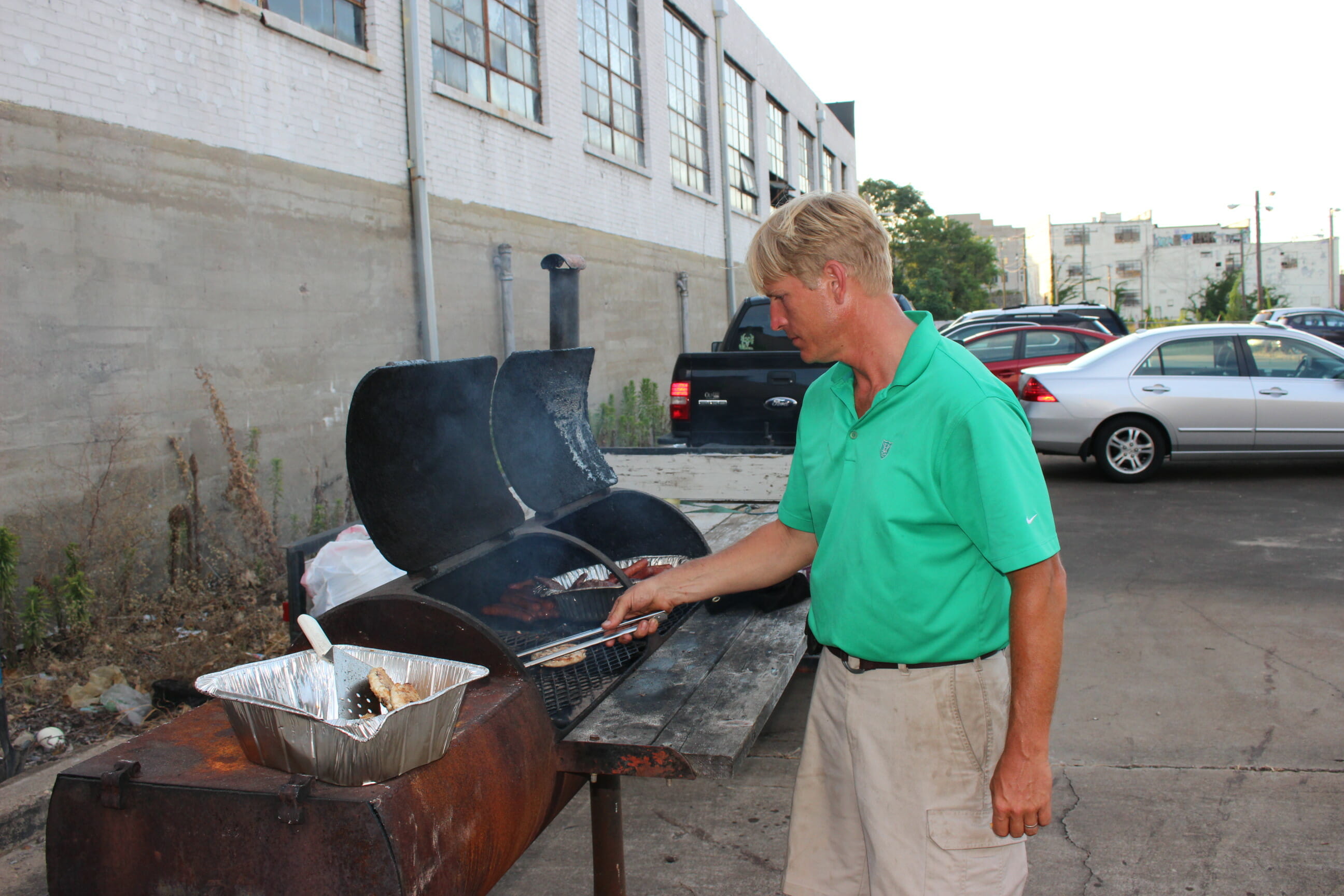 a man wearing khaki shorts and a green polo, grilling in a parking lot.